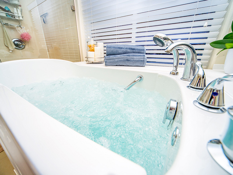 The image shows a modern bathroom with a freestanding bathtub filled with water, featuring chrome fixtures and a blue-toned color scheme.
