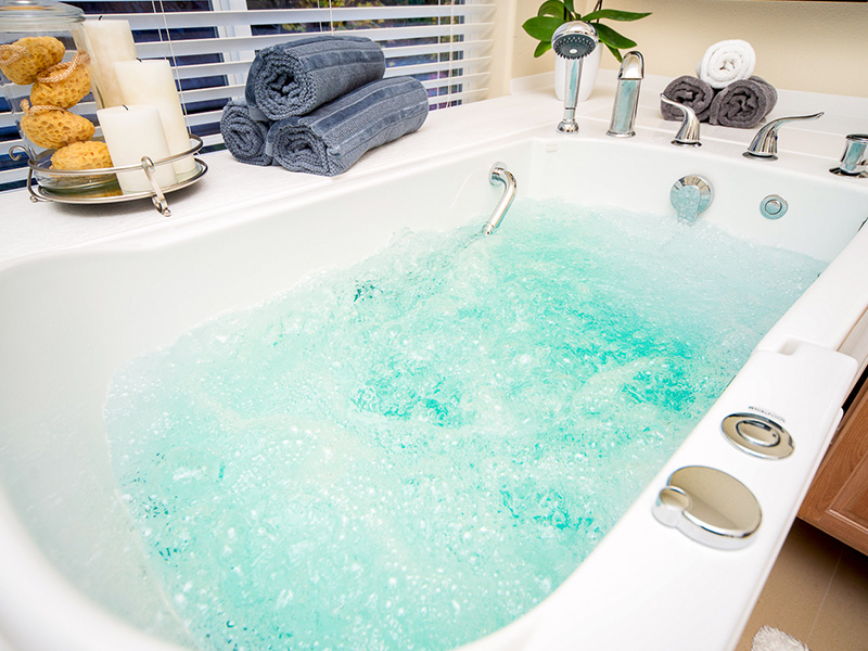 The image shows a bathroom with a large bathtub filled with bubbles, surrounded by towels on a rack and a bottle of bath products, situated next to a window with blinds partially drawn.