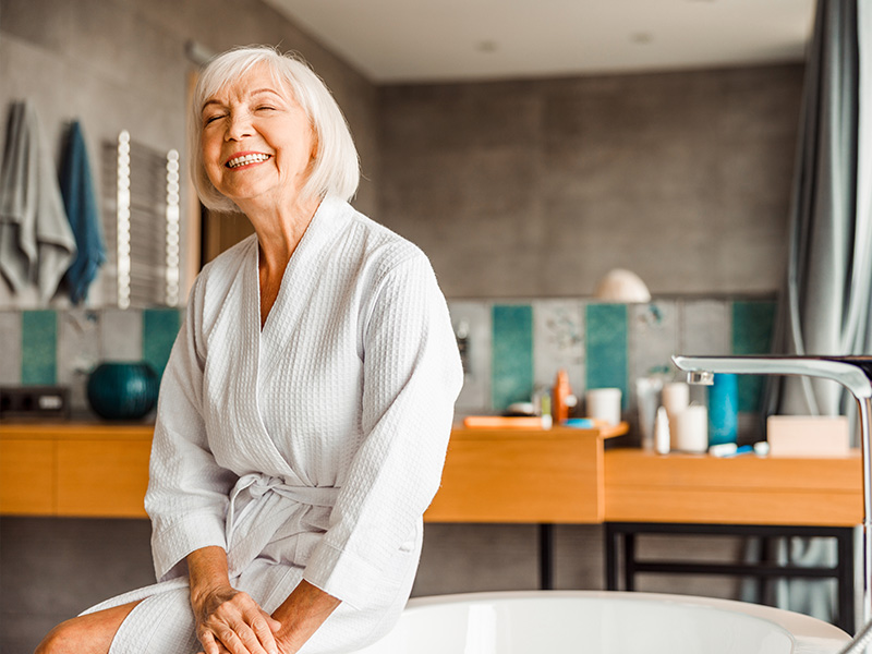 A woman wearing a robe sits on the edge of a bathtub with a smile, looking towards the camera.