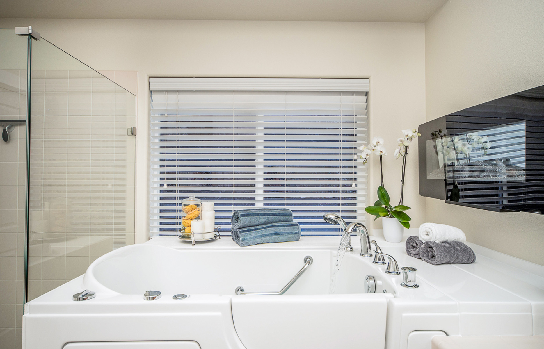 The image shows a modern bathroom interior with a large white bathtub, a glass-enclosed shower area, a double sink vanity, a mirror over the vanity, a black cabinet, and a window with blinds allowing natural light into the room.