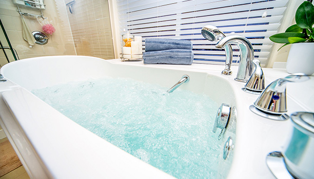 The image shows a modern bathroom with a large soaking tub filled with water, featuring a faucet and two handles for temperature control, placed next to a window with blinds partially drawn.