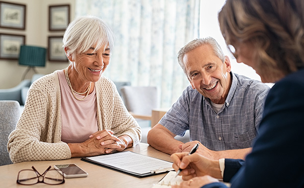 The image shows two elderly individuals sitting at a table with papers, while another person is standing across from them, smiling and interacting with them.