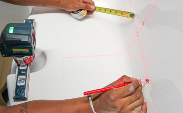 The image shows three individuals engaged in an activity involving a whiteboard with red lines drawn on it, where they are using markers to trace the outline of the lines, suggesting a collaborative task related to planning or design.
