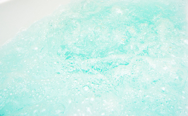 The image displays a close-up view of a bright green crystalline substance with a speckled texture, resembling a gemstone or a type of mineral, set against a blurred background that suggests a natural outdoor setting.