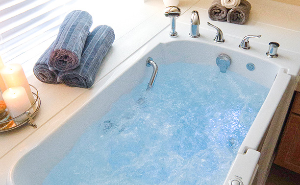 The image shows a luxurious bathtub filled with blue water, placed in a bathroom setting, with towels neatly rolled on the edge of the tub, and a candle holder with a lit candle nearby.