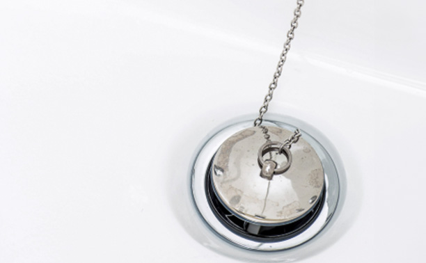 The image shows a small, round silver-colored pendant with a unique design featuring a coin-like shape and a chain attached to a small ring at the top, displayed against a white background with a reflective surface beneath it.